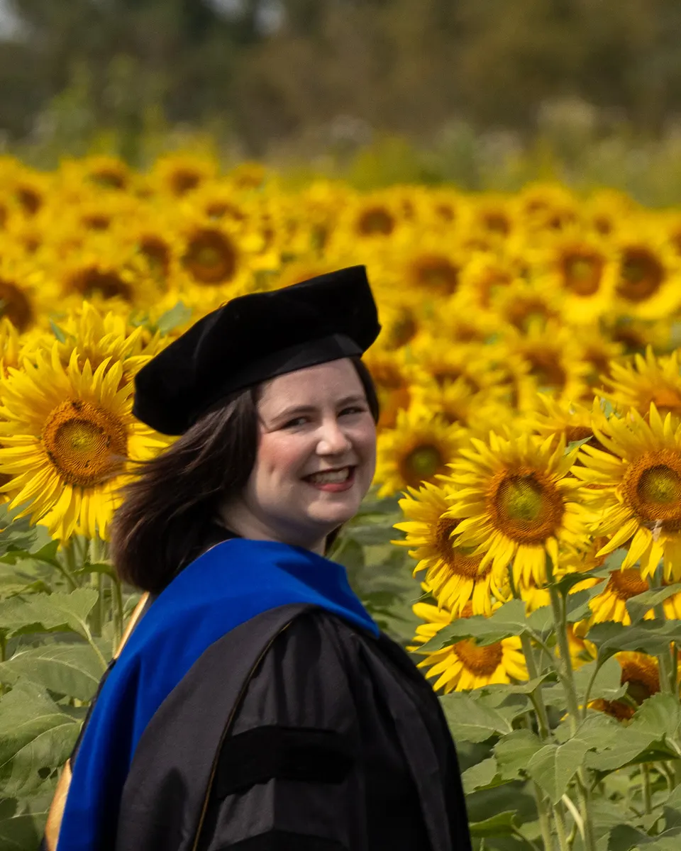 Sadie Nickles in her doctoral gown and hood, smiling over her shoulder in a field of sunflowers
